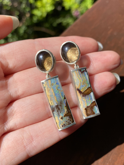 hand holding a pair of earrings with round smokey quartz and rectangular boulder opal drop