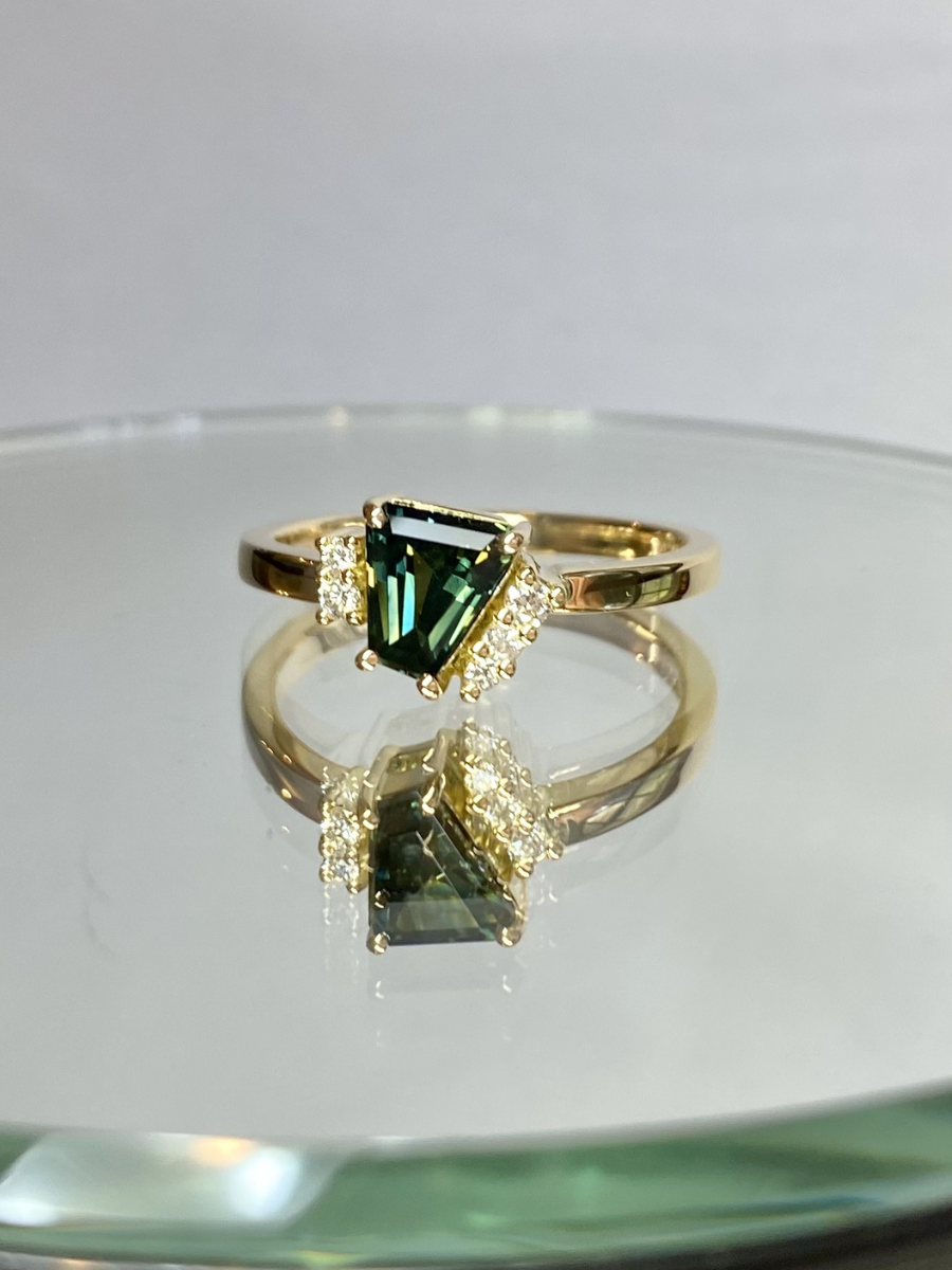 Gold ring with a teal sapphire and accent diamonds on a reflective surface