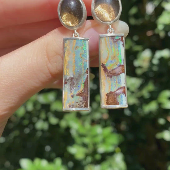 pair of earrings with round smokey quartz and rectangular boulder opal drop held in a hand moving in the sinlight
