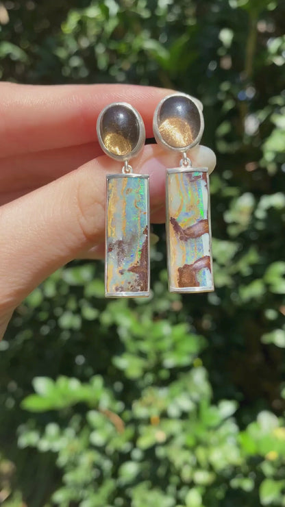 pair of earrings with round smokey quartz and rectangular boulder opal drop held in a hand moving in the sinlight